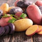 Verschiedene ganze und geschnittene Kartoffeln in lila, gelb und rot auf einer Holzoberfläche, garniert mit Petersilieblättern.