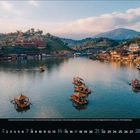 11 mit kleinen Zahlen darunter. Traditionelle Boote auf einem ruhigen See, umgeben von Hügeln mit Gebäuden bei Sonnenuntergang.