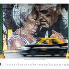 Unter einem großen Wandgemälde eines älteren Paares fährt ein schwarz-gelbes Taxi entlang. Darunter ein Kalender mit November.