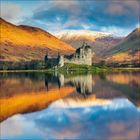 Keine erkennbaren Texte. Eine Burgruine spiegelt sich in einem ruhigen See, umgeben von herbstlich gefärbten Hügeln.