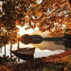 Ein idyllischer See im Sonnenuntergang, ein Boot am Ufer, umgeben von herbstlichem Laub und Bergen im Hintergrund.