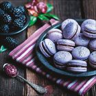 Lila Macarons auf einem Teller, daneben Brombeeren in einer Schale und ein Löffel mit Marmelade auf einem gestreiften Tuch.