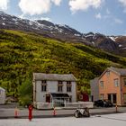 Ein Ortsschild mit "San Kafe". Mehrere kleine Häuser vor einem bewaldeten Berg, blauer Himmel mit wenigen Wolken.