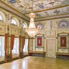 Ein prunkvoller Raum mit dekorativer Decke, großen Fenstern mit Vorhängen, Statuen an den Wänden und einem Kronleuchter.