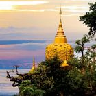 Ein goldener buddhistischer Stupa auf einem Felsen, umgeben von grünen Bäumen, vor einem farbenfrohen Sonnenuntergangshimmel.