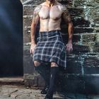 Text: "November". Ein tätowierter Mann trägt einen karierten Kilt, steht vor einem steinernen Hintergrund und blickt selbstbewusst.