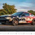 Text: November, 2014er Ford Mustang. Auto mit US-Flaggen-Design, schwarzen Felgen, auf einer Straße vor Bäumen bei klarem Himmel.