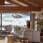 Keine erkennbaren Texte. Moderner Raum mit hellem Sofa, Holzdecke, großen Fenstern; Blick auf schneebedeckte Landschaft.