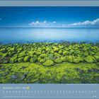 November 2027 Kalender mit grünen, algenbedeckten Steinen am Ufer, dahinter ruhige See und blauer Himmel mit Wolken.
