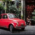 "Merci" auf Nummernschild, Kalender unten, Fiat 500 beschriftet. Roter Oldtimer vor Café, Frau am Tisch, viele Bücher.