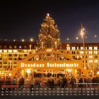 "Dresdner Striezelmarkt" leuchtet auf einem Weihnachtsmarkt. Ein großer, beleuchteter Baum steht im Hintergrund.