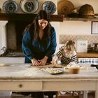 Text: „BREAD“. Eine Frau und ein Kind machen Teig auf einem Küchentisch. Teller und Körbe im Hintergrund.
