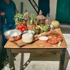 Ein Holztisch mit Käse, Gemüse, Kräutern, Tomaten, Artischocken und Gewürzen. Zwei Personen im Hintergrund.
