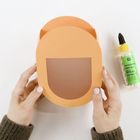 Bastelkleber Hobby Glue. Zwei Hände halten einen orangenen Papierhelm; rechts steht eine Flasche Kleber.