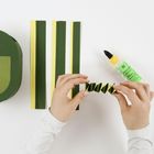 Zwei Hände falten grünes Papier; daneben liegen lange Papierstreifen in Grün und Gelb und eine Flasche Bastelkleber.