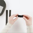 Keine erkennbaren Texte. Zwei Hände falten schwarzen Papierstreifen in Ziehharmonikaform. Ein dickerer, runder Karton links.
