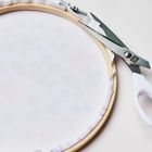 Eine Schere mit weißem Griff und einem Stickrahmen, der straff gespannt ist. Der Stoff zeigt ein blasses Blumendesign.