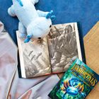 Text: "The Last Immortals: Dawn of the Axolotl." Ein Plüschtier liegt auf einem Buch mit einer Schwarz-Weiß-Illustration.