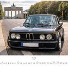 BMW 323i. Kalender mit hervorgehobenem Tag 12. Vordergrund: schwarzes Auto. Hintergrund: Brandenburger Tor, Bäume.