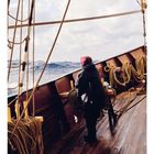 Nummer 12 und Kalendertage. Eine Person mit rotem Kopftuch steht auf einem Holzdeck eines Segelschiffs, umgeben von Seilen.