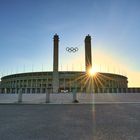 Im Vordergrund stehen zwei Säulen mit den olympischen Ringen; dahinter das Olympiastadion bei Sonnenuntergang.