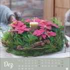 "Dez" in weißer Schrift oben links. Adventskranz mit roten Blüten und Kerze auf Holztisch. Kalender mit Wochentagen darunter.