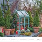 Text: Dezember. Szene mit kleinen Tannenbäumen, Kerzen und einem türkisfarbenen Glashaus auf einem Holztisch.