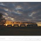 DEZEMBER. Abendsilhouette: Zug bei Sonnenuntergang, Dampfwolken, Baum im Vordergrund, dramatischer wolkiger Himmel.