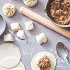 Teigstücke, Nudelholz und gefüllte Teigtaschen auf mehlbedeckter Oberfläche. Eine Schüssel mit Füllung und ein Löffel.