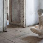 Keine Texte zu erkennen. Eine Skulptur einer kauernden Person in einem alten, leeren Raum mit Holzboden und weißen Wänden.