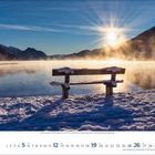 Kalenderseite für Dezember 2023. Eine schneebedeckte Bank am ruhigen See, umgeben von Bergen, Sonnenaufgang im Hintergrund.