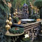 Goldene Dekorationen, Kerzen, immergrüne Kränze und eine Engelsstatue in einem üppig grünen, festlichen Garten.
