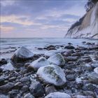 Steinige Küste mit schneebedeckten Felsen, dahinter das Meer unter einem bewölkten Himmel bei Sonnenuntergang.