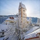 Ich sehe keine Texte. Ein Kirchturm ragt aus einer schneebedeckten Winterlandschaft hervor, die Sonne strahlt dahinter.