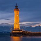 Ein beleuchteter Leuchtturm mit Uhr auf einer Mole, umgeben von Wasser und Bergen im Hintergrund bei Abenddämmerung.