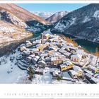 Dezemberkalenderseite mit verschneitem Dorf in Berglandschaft; Fluss und Brücke, umgeben von schneebedeckten Hügeln.
