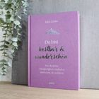Text vorne: "Kira Geiss. Du bist kostbar & wunderschön. Wie du deine Einzigartigkeit entdeckst und lernst, sie zu feiern."  
Ein lila Buch steht auf einem Holztisch, links daneben Pflanzendekoration.