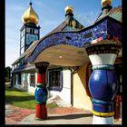 XII 2027. Buntes Gebäude mit goldenen Kuppeln und säulenartiger Struktur im Stil von Hundertwasser.