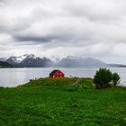 Rotes Haus auf grüner Wiese am Wasser, im Hintergrund Berge mit Schneekappen unter bewölktem Himmel.
