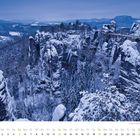 Dezemberkalender: Schnee bedeckte Felsen und Bäume in einer winterlichen Landschaft bei Morgendämmerung.