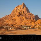 Namibia 11. Spitzkoppe, Damaraland. Drei Hütten vor einem großen, felsigen Berg im Wüstenlicht.