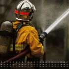 Kalender mit Tagen und Daten. Ein Feuerwehrmann in gelber Uniform spritzt Wasser mit einem Schlauch. Nummer 12 sichtbar.