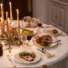 Ein festlich gedeckter Tisch mit Kerzenleuchtern, Fleisch, Kartoffeln, Brot, Salat und Weingläsern auf einer weißen Tischdecke.