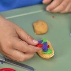 Hände formen bunte Knetmasse auf einem Tisch; Behälter und Spielzeugmesser in der Nähe.