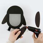 Eine Hand hält ein schwarzes Pappteil, während eine Heißklebepistole Kleber darauf aufträgt. Im Hintergrund Pinguin-Bastelmaterial.