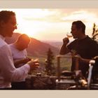 Vier Männer genießen Speisen und Getränke im Freien bei Sonnenuntergang, mit Bergen und Bäumen im Hintergrund.