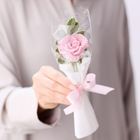 Keine Texte im Bild. Jemand hält eine gehäkelte rosa Rose mit grünen Blättern, verpackt in Plastik, gebunden mit rosa Schleife.