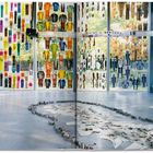 Dieses Kunstwerk zeigt bunte Kleidung, die an großen Fenstern hängt. Auf dem Boden liegen Papiere, umrandet von Steinen.