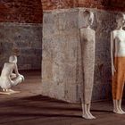 Drei skulpturale Figuren vor einer grob gemauerten Ziegel- und Steinwand in einem weiten Raum.