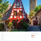 09. Woche, März 2027. Ein rot-weißes Fachwerkhaus, daneben ein steinerner Turm, idyllisch mit Bäumen. Glasklarer blauer Himmel.